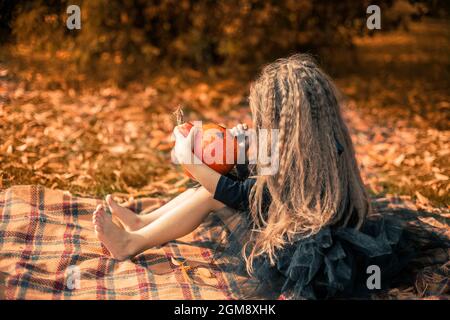 Halloween. Mädchen mit langen Haaren in schwarzem Kleid sitzt auf einer orangefarbenen Decke im Park und zieht ein Gesicht auf einen Kürbis. Stockfoto