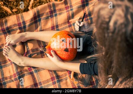 Halloween. Mädchen mit langen Haaren in schwarzem Kleid sitzt auf einer orangefarbenen Decke im Park und zieht ein Gesicht auf einen Kürbis. Stockfoto