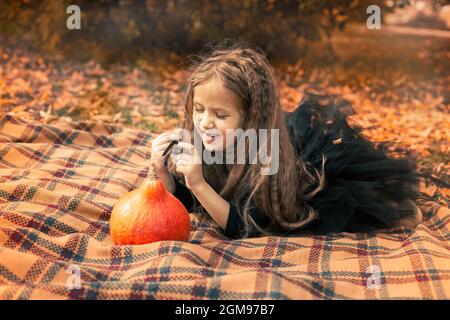 Halloween. Mädchen mit langen Haaren in schwarzem Kleid sitzt auf einer orangefarbenen Decke im Park und zieht ein Gesicht auf einen Kürbis. Stockfoto