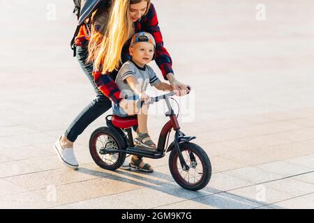 Das kleine glückliche Kind lernt mit ihrer Mutter im Park im Park Fahrrad zu fahren, die glückliche junge Mutter lehrt ihren Sohn, im Sommer Fahrrad im Park zu fahren Stockfoto