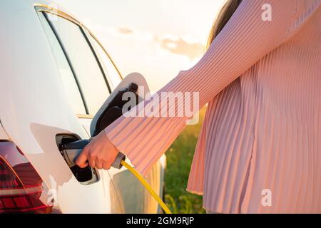 Frau lädt bei Sonnenuntergang ein weißes Elektroauto an einer Ladestation im Naturfeld nahe der Autobahn Stockfoto