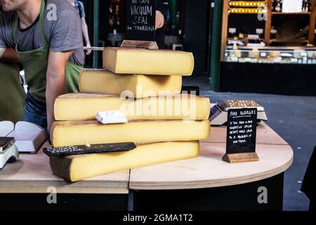 Große Käseplatten auf einem Holztisch auf einem Markt, auf dem sich der Arbeiter über das Gespräch mit dem Kunden lehnte, und auf Tischen mit Waage und Salbe Schilder für Lebensmittelallergien Stockfoto