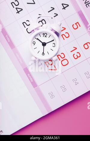 Weißer Wecker und Kalender auf einem rosa Desktop-Hintergrund. Frauentage Stockfoto