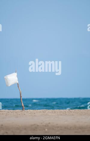 Menschenleerer Strand mit einer weißen Flagge, die mit einem Holzstab im Sand und einer Plastiktüte, die vom Wind mit dem Meer und dem blauen Himmel geblasen wird, versehen ist. Selecti Stockfoto