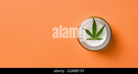 Hanf Hautpflege Feuchtigkeitscreme und grünes Cannabis Sativa Blatt in einem Glas auf hellorangefarbenem Hintergrund. Kräuterkosmetik mit medizinischem Marihuana. Stockfoto