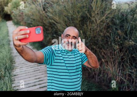 Ein glücklicher Mann mittleren Alters zeigt den Daumen hoch, während er ein Selfie mit einem Mobiltelefon im Freien in der Natur macht. Stockfoto
