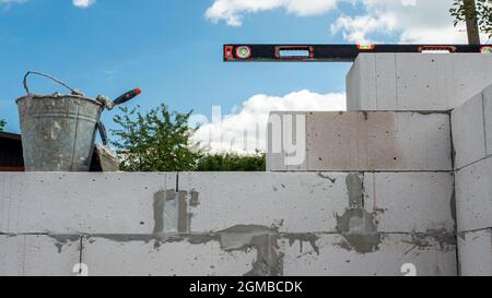 Ein Haus aus einem weißen Block bauen, Werkzeuge auf einem Ziegelstein bauen. Gebäudehüfe mit Flüssigkeit, Eimer mit Spachtel und Zement Stockfoto