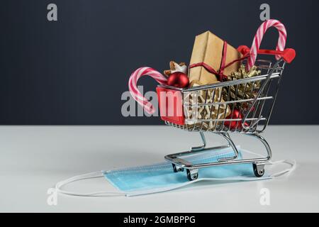 Kleiner Warenkorb mit Geschenk, weihnachtsdekoration und Süßigkeiten auf einer chirurgischen Gesichtsmaske gegen Covid-19-Infektion, sichere Ferienkäufe während der c Stockfoto