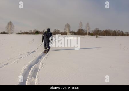 Ein Jäger auf breiten Skiern geht mit einer Waffe über ein schneebedecktes Feld und wirft einen langen Schatten. Von der Jagd zurückkehrend, geht er zu den Häusern im d Stockfoto