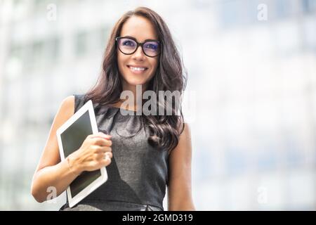 Lächelnde Frau hält lächelnd ein Tablet und geht in Geschäftskleidung vor das Bürogebäude. Stockfoto