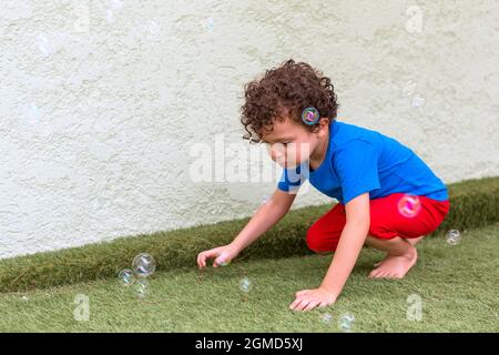 Schöner 4-5-jähriger kaukasischer Junge mit lockigem Haar in seinem Hinterhof, der mit Seifenblasen spielt Stockfoto