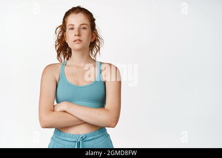 Junge selbstbewusste Rothaarige Frau kreuzt die Arme auf der Brust und sieht motiviert aus. Joggingweibchen beim Sport, Training im Fitnessstudio mit aktivem Ohr, weißer Hintergrund Stockfoto