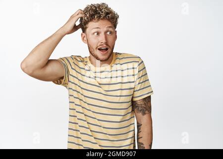 Überraschte junge blonde Mann, der schockiert auf Banner, kratzenden Kopf, stehend in T-Shirt vor weißem Hintergrund sah Stockfoto