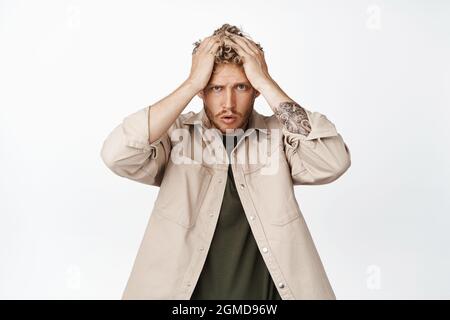 Frustrierter junger Mann in Schwierigkeiten, die Hände am Kopf haltend, bekümmert und verärgert, großes Problem habend, vor weißem Hintergrund in Verzweiflung stehend Stockfoto