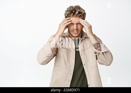 Der junge blonde Mann berührt seinen Kopf und grimazt vor Schmerzen, hat starke Kopfschmerzen, steht mit Migräne oder Kater vor weißem Hintergrund Stockfoto