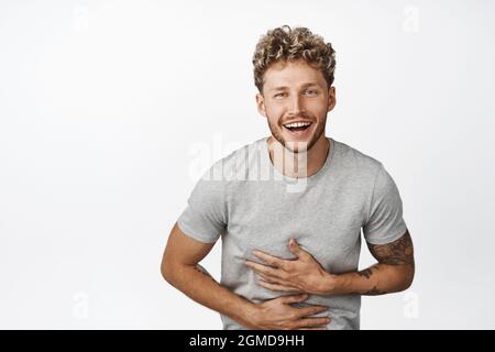 Fröhlicher, ehrlicher Kerl, der lachend und lächelnd etwas Lustiges ansah, mit Händen, die die Brust berühren, auf weißem Hintergrund stehend Stockfoto