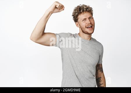 Motivierter blonder Kerl protestiert, Faust hochheben und rufen, smetschen, Bizeps beugen und mit seinem fetten und sportlichen Körper prahlen, in dem er steht Stockfoto