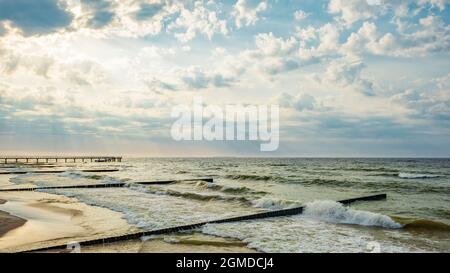 Wellige Ostsee vor Sonnenuntergang, Zelenogradsk Pier und Wellenbrecher in der Nähe des Ufers, Sonnenstrahlen brechen durch die Wolken Stockfoto