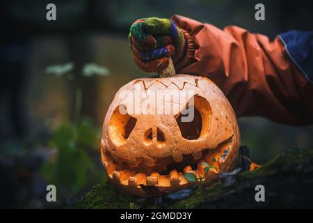 Hand, die den Deckel von Jack O Lantern entfernt, mit einem bösen Gesicht. Geschnitzter Kürbis für Halloween auf gefallenen Bäumen im Nachtwald Stockfoto