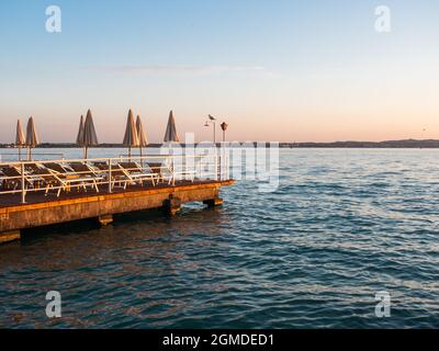 Badeplattform oder Terrasse mit Sonnenschirme und Strandliegen am Gardasee in Sirmione, Italien an einem Sommerabend Stockfoto