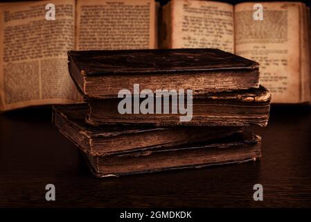 Stapel alter abgenutzter, schäbiger jüdischer Bücher in Ledereinband und offener, verschwommener Tora im Hintergrund im Dunkeln. Nahaufnahme. Selektiver Fokus Stockfoto