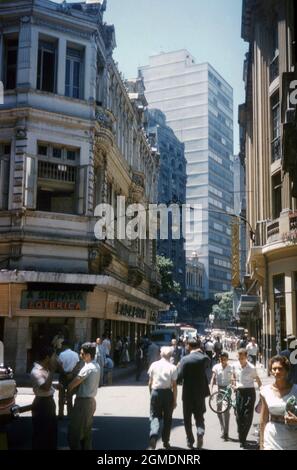 Eine geschäftige Seitenstraßenszene im Sonnenschein im Zentrum von Rio de Janeiro, Brasilien, um 1959. Die Architektur ist eine Mischung aus dem alten Kolonialstil und modernen Gebäuden. Schilder werben (auf Portugiesisch) für eine Lotterie-Verkaufsstelle (links) und ein Geschäft, das Industrieschuhe verkauft (rechts). Dieses Bild stammt von einem alten Kodak-Amateurfotograf mit Farbtransparenz – einem Vintage-Foto aus den 1950er Jahren. Stockfoto