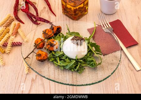 Gastronomische Komposition aus Burrata-Käseplatte mit gegrillten Kirschtomaten auf einem Rucola-Bett mit Gabel und Serviette im Hintergrund Stockfoto