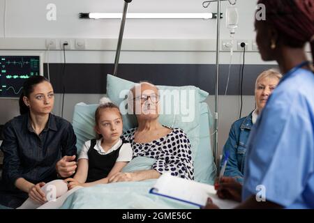afroamerikanische Krankenschwester, die mit einem älteren älteren Mann auf der Krankenstation über die Behandlung von Krankheiten spricht. Eine fröhliche, fürsorgliche Familie, die neben einem kranken Patienten sitzt, unterstützt ihn während der klinischen Genesung Stockfoto