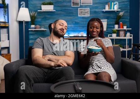 POV von interracial paar genießen Aktivität zusammen zu Hause. Gemischte Rennpartner lächeln und sehen im Wohnzimmer Fernsehen, schauen auf die Kamera und essen Popcorn-Snack im Fernsehen Stockfoto
