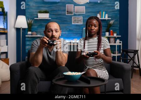 POV von interracial paar spielen Videospiel auf Konsole hält Controller im Fernsehen. Multiethnische Partner blicken auf die Kamera und genießen lustige Aktivitäten mit TV und Joysticks Stockfoto