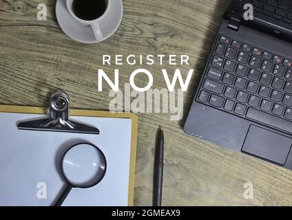 Draufsicht des Textes JETZT REGISTRIEREN, Kaffee, Laptop, Stift und Lupe. Stockfoto