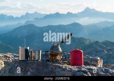 Morgenkaffee mit Aussicht. Kaffeekocher und Tassen voller heißer Getränke auf dem Gasbrenner hoch in den Bergen. Outdoor-Paradies und Fernweh-Konzept Stockfoto