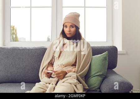 Verärgert Frau mit Fieber in warmen Strickschal und Hut sitzen auf dem Sofa mit Thermometer in den Händen. Stockfoto