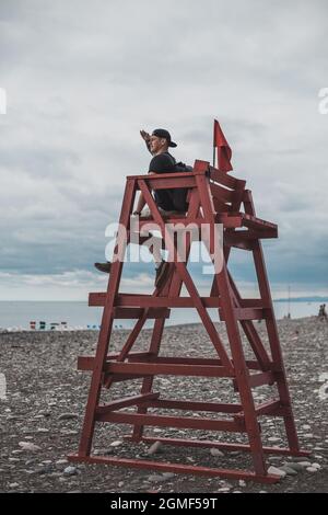 Der Mann sitzt auf einem Rettungsturm mit der roten Flagge am dunklen Kiesstrand in Batumi, Georgia. Blick auf das Meer. Hochwertige Fotos Stockfoto