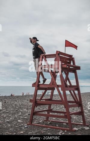 Der Mensch steht auf einem Rettungsturm mit der roten Flagge am dunklen Kiesstrand in Batumi, Georgia. Blick auf das Meer. Hochwertige Fotos Stockfoto