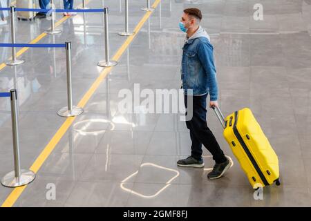 Mann mit medizinischer Gesichtsmaske vor dem Check-in-Schalter im Flughafeninneren während der Zwangsabsperrung. Urbane Freizeitreisende mit Kofferraumbeförderung Stockfoto