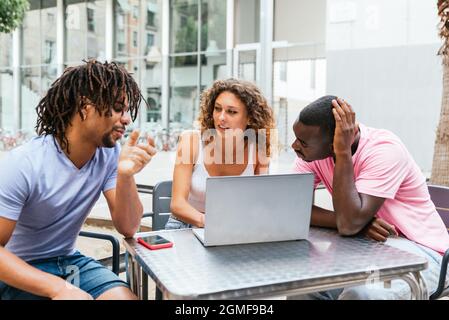 Junge kaukasische Frau, die draußen auf einem Tisch mit Laptop sitzt und einen lateinischen Mann anschaut. Konzept der Muti ethnischen Gruppe von Studenten Stockfoto