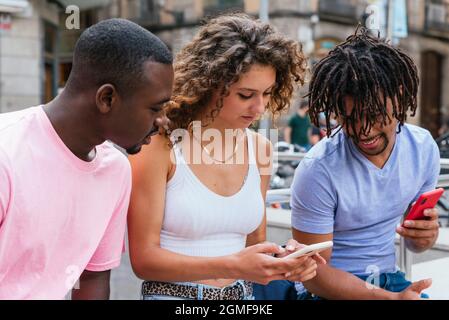kaukasische junge Frau, die ihren Telefonbildschirm afrikanischen und lateinischen männlichen Freunden zeigt Stockfoto