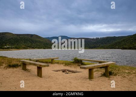 Vier Holzbänke an einem Strand mit Wasser und Hügeln im Hintergrund. Stockfoto