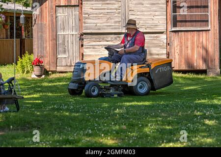 Velena, Lettland - 19. Juni 2021: Ein älterer Mann mit einem Rasenmäher mäht das Gras auf dem Hof des Landhauses Stockfoto
