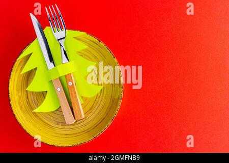 Weihnachtsbesteck. Weihnachtliche Tischeinstellung auf rotem Hintergrund mit gelbem Teller, Besteck und Papierweihnachtsbaum. Draufsicht Stockfoto