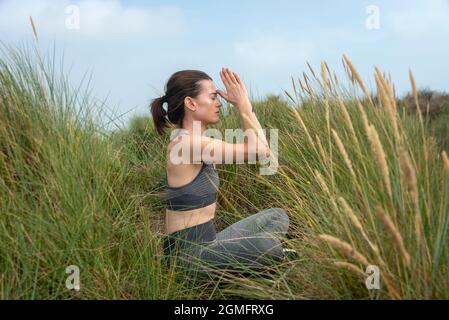 Seitenansicht einer in den Dünen sitzenden Frau, die meditiert und Yoga praktiziert. Gesundes Lifestyle-Konzept. Stockfoto