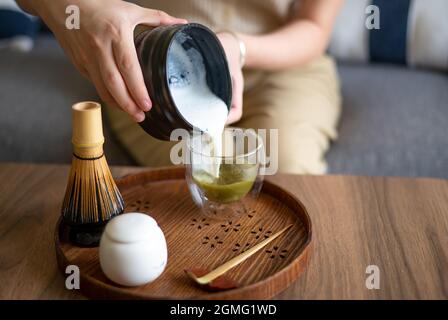 Frau, die zu Hause mit traditionellem Bambusset für Teezeremonien aufgeschäumte Milch in ein fertiges japanisches Matcha-Getränk gießt, während sie auf dem Sofa A sitzt Stockfoto