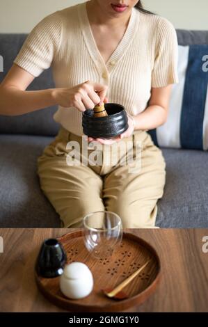 Frau, die zu Hause auf dem Sofa sitzt und japanische Matcha-Getränke mit traditionellem Bambus-Set und natürlichem Schneebesen mischt. Stockfoto