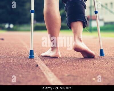 Sportlerin mit Krücken, Handgelenkstütze und Kniestütze, gebändertes Bein. Frau läuft auf roter Lauffläche Stockfoto