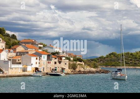 Prvic Luka, Kroatien - 25. August 2021: Steinhäuser mit Sonnenschirmen und Stühlen vor der Tür und Boote an der Küste gebunden und ein Segelboot im Meer Stockfoto