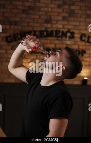 RIVNE, UKRAINE JUNI 26 2020: Junger bärtiger Mann trinkt Coca Cola aus der Flasche im Café auf Backsteinmauer Hintergrund. Stockfoto