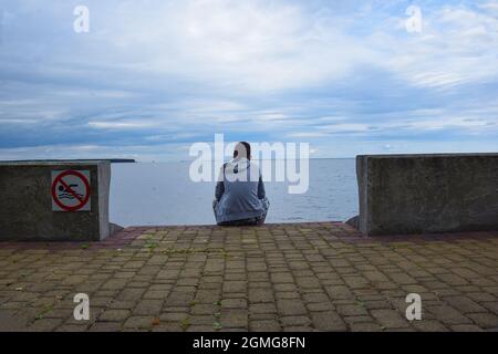 Ein junger Mann sitzt mit dem Rücken auf dem Damm, an der Küste. Der Himmel ist düster. Links Verbotsschild zum Schwimmen Stockfoto