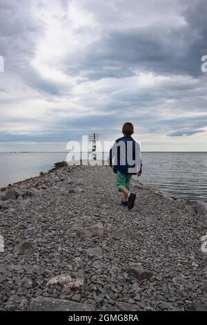 Ein Junge mit dem Rücken drehte sich und ging auf einen fernen Leuchtturm entlang eines Steges aus Steinen. Der Himmel ist mit Wolken bedeckt Stockfoto