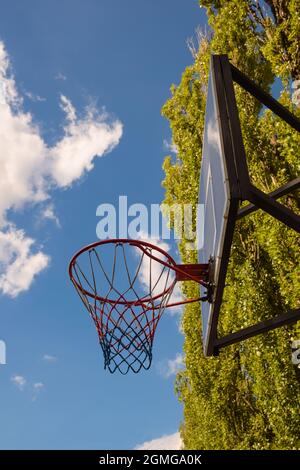 Basketball-Korb und Backboard gegen den blauen Himmel mit Wolken und Bäumen. Fotografiert von unter dem Schild, von unten Stockfoto